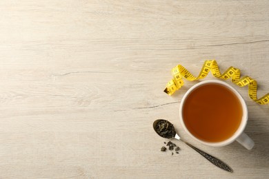 Cup of herbal diet tea and measuring tape on light wooden table, flat lay with space for text. Weight loss concept