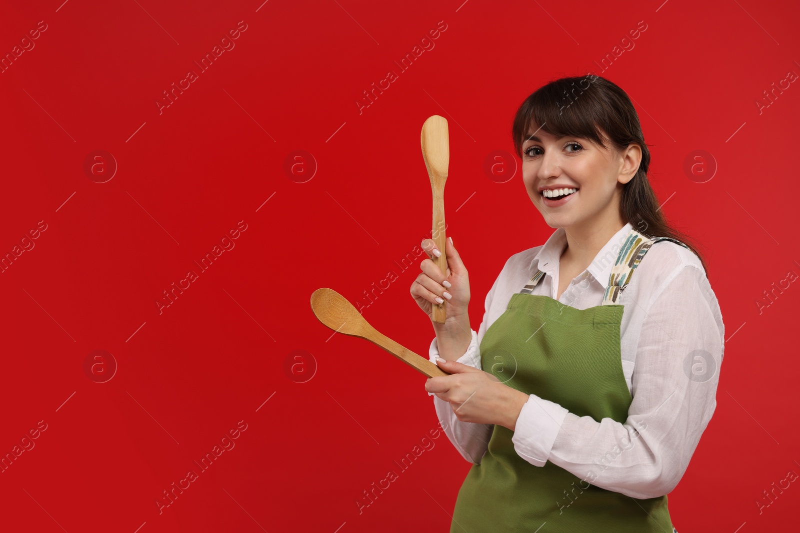 Photo of Happy professional confectioner in apron holding wooden spoon and spatula on red background. Space for text