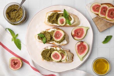 Tasty bruschettas with cream cheese, pesto sauce, figs and fresh basil on white tiled table, flat lay