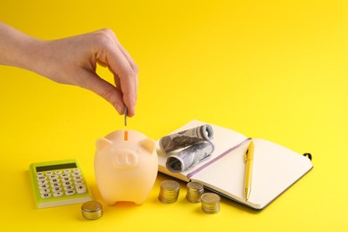 Financial savings. Woman putting coin into piggy bank on yellow background, closeup