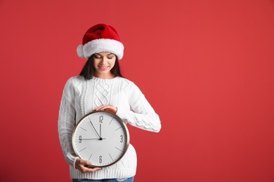 Young beautiful woman in Santa hat holding big clock on color background. Christmas celebration