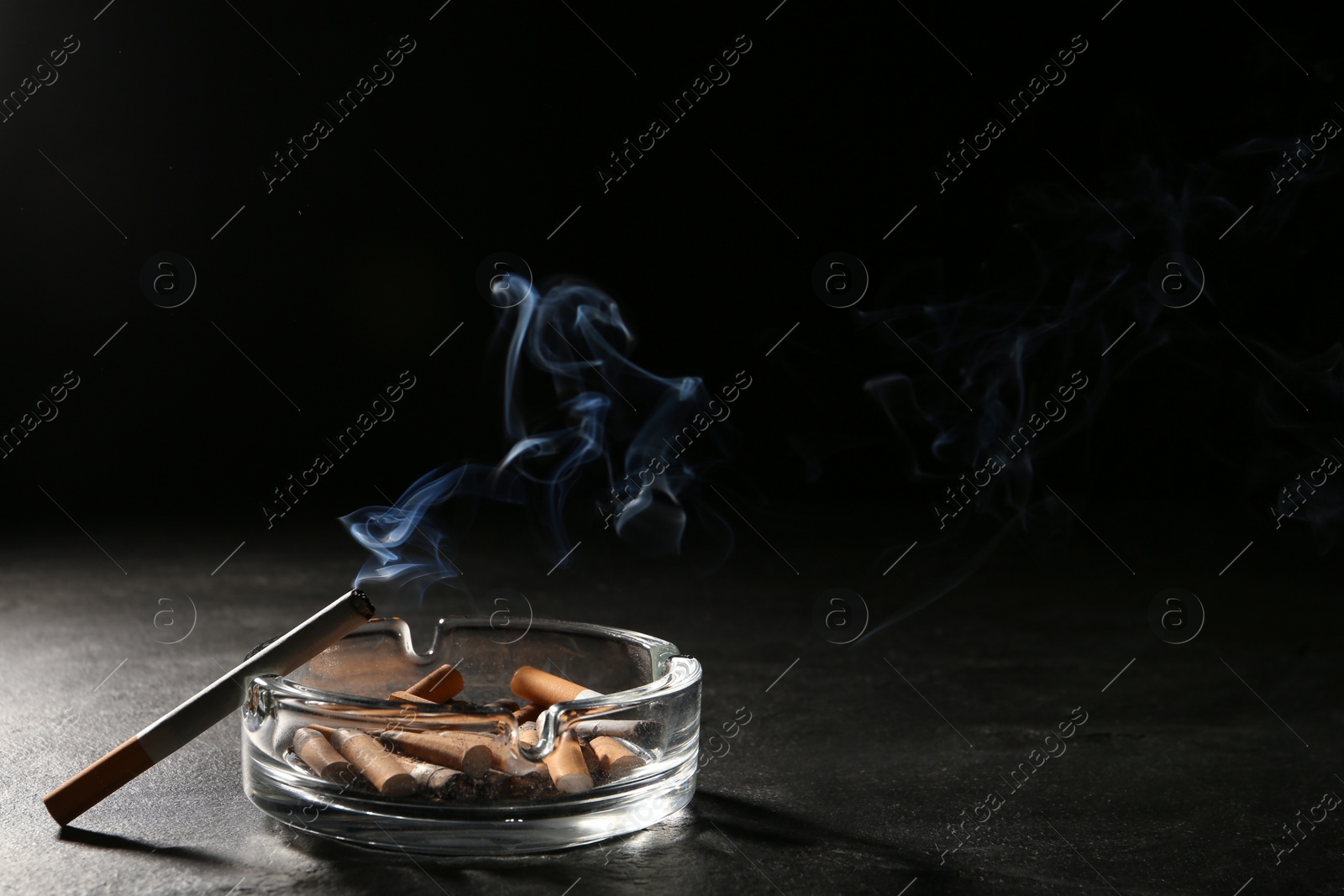 Photo of Glass ashtray with stubs and smoldering cigarette on grey table against black background. Space for text