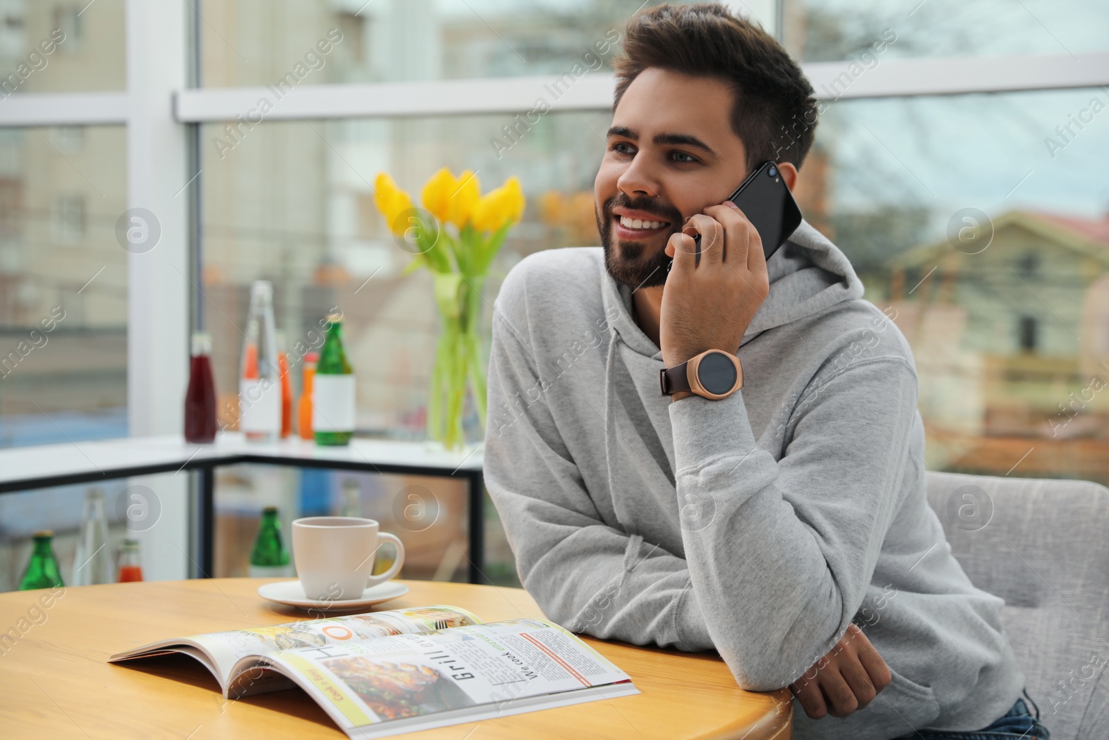 Photo of Happy young man wearing smart watch and talking on smartphone at table in cafe