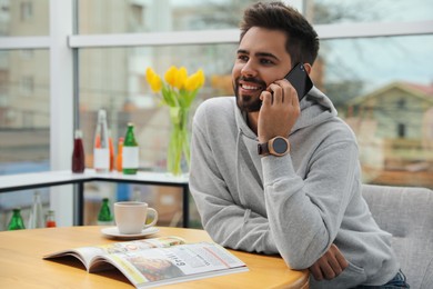 Photo of Happy young man wearing smart watch and talking on smartphone at table in cafe