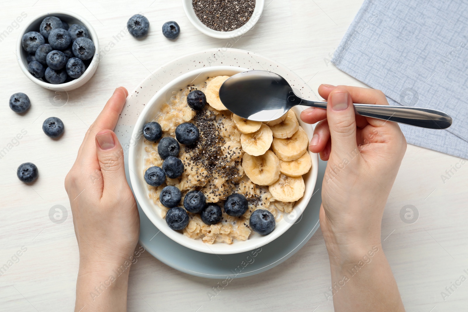 Photo of Woman eating tasty oatmeal with banana, blueberries and chia seeds at white wooden table, top view