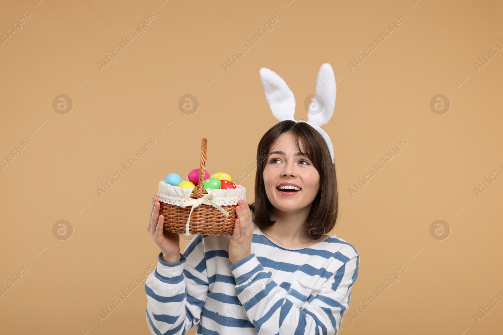 Photo of Easter celebration. Happy woman with bunny ears and wicker basket full of painted eggs on beige background