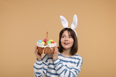 Photo of Easter celebration. Happy woman with bunny ears and wicker basket full of painted eggs on beige background