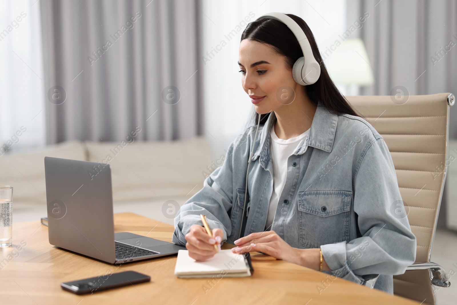 Photo of Young woman in headphones watching webinar at table in room