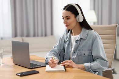 Photo of Young woman in headphones watching webinar at table in room