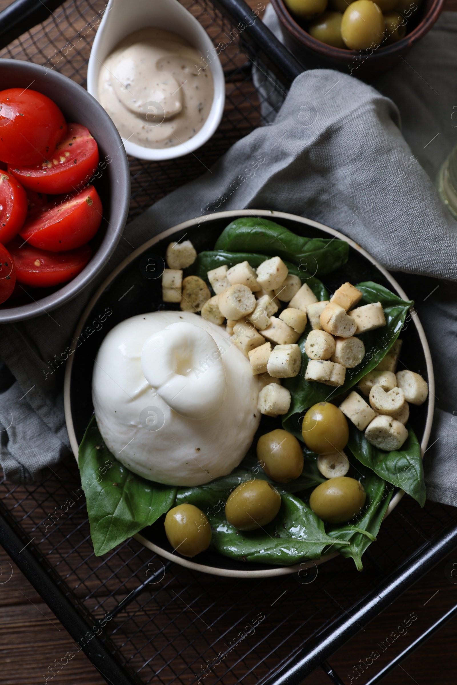 Photo of Delicious burrata cheese served with olives, croutons, basil and tomatoes on wooden table, top view