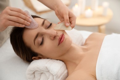 Photo of Young woman receiving facial massage with gua sha tools in beauty salon