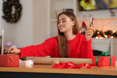 Photo of Beautiful young woman with scissors decorating Christmas gift at table in room