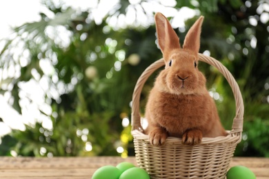 Photo of Cute bunny, basket and Easter eggs on table against blurred background. Space for text