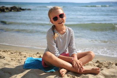 Happy little girl in stylish sunglasses on sandy beach near sea