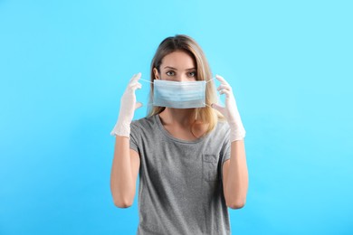 Woman in medical gloves putting on protective face mask against light blue background
