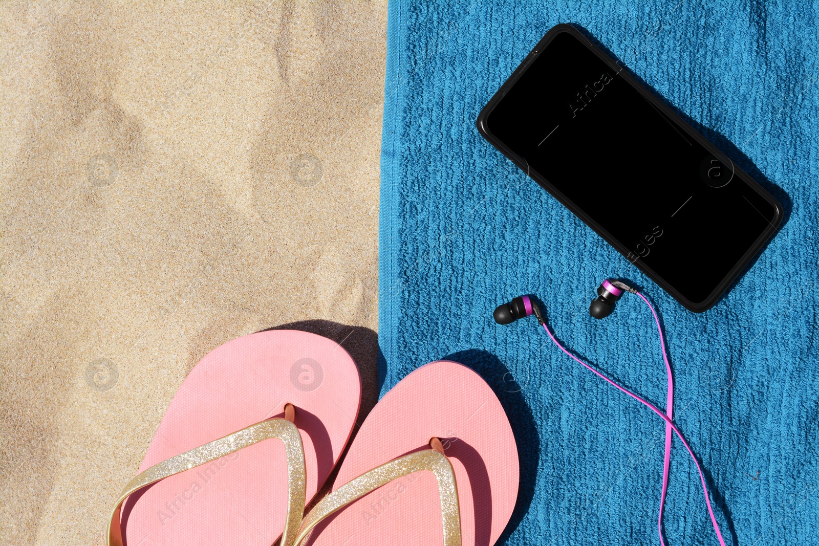 Photo of Soft blue beach towel with flip flops, smartphone and earphones on sand, flat lay. Space for text