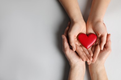 Young and elderly women holding red heart on light grey background, top view. Space for text