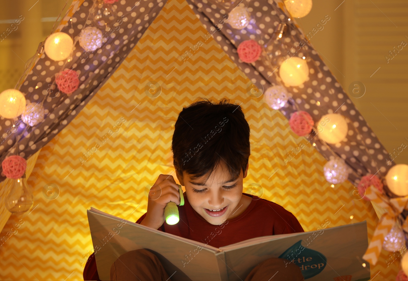 Photo of Boy with flashlight reading book in play tent at home