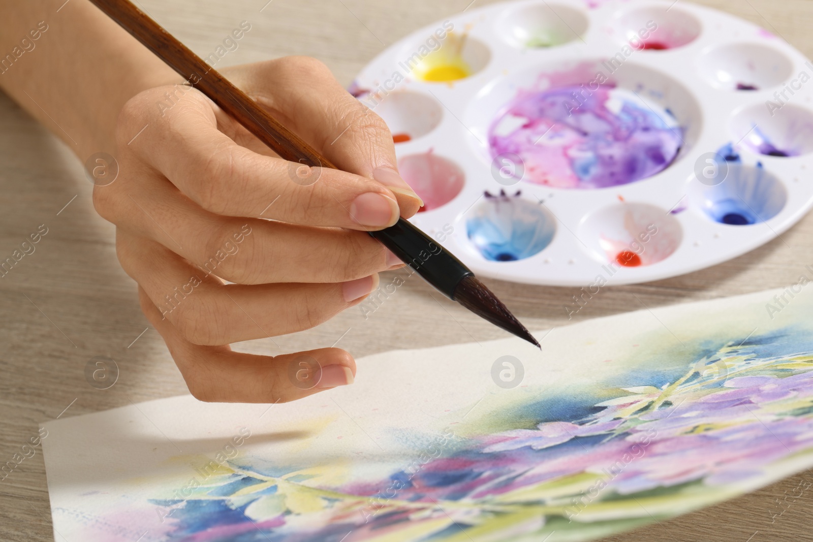 Photo of Woman painting flowers with watercolor at white wooden table, closeup. Creative artwork