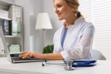 Photo of Doctor with laptop at white table, focus on stethoscope. Patient consultation