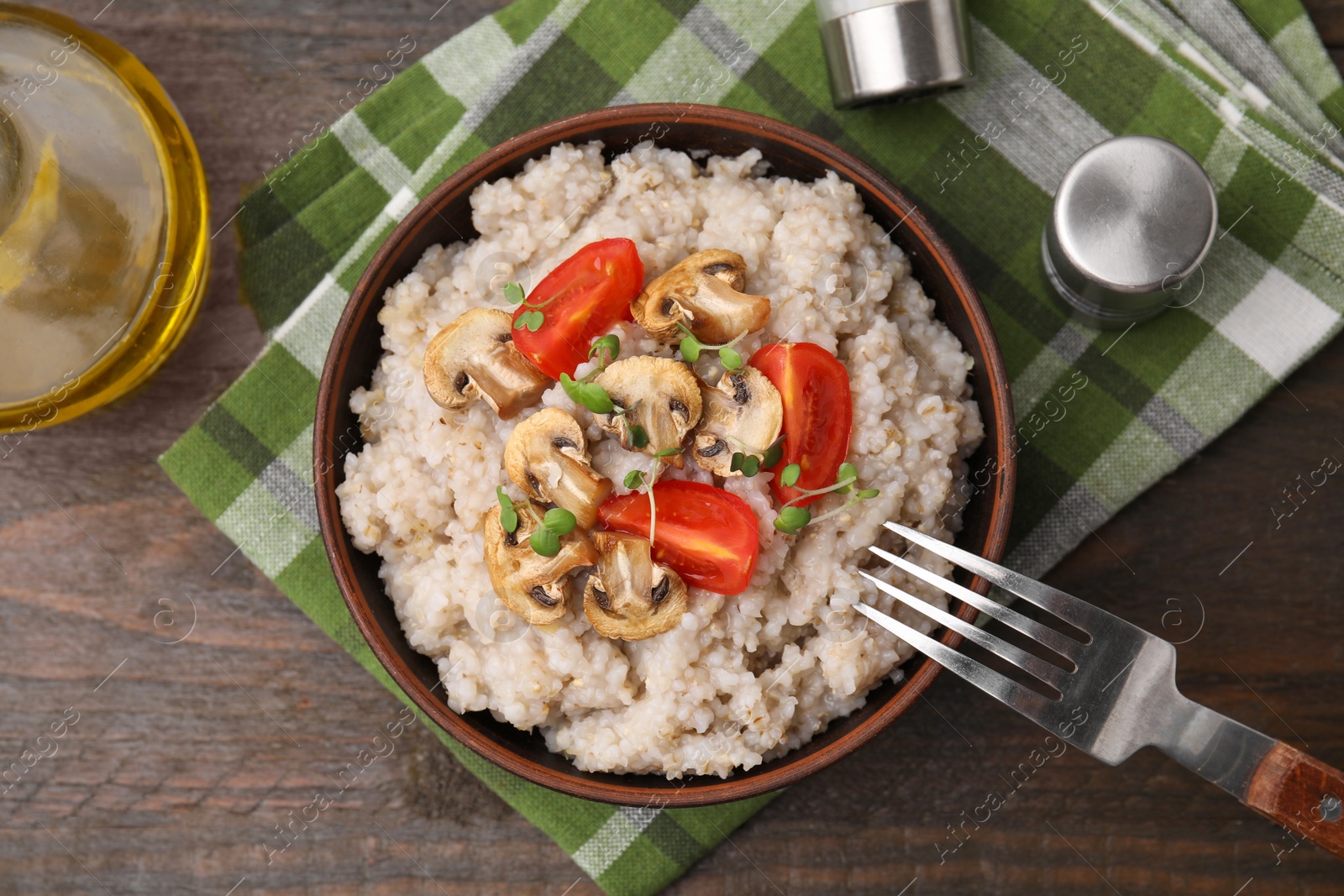 Photo of Delicious barley porridge with mushrooms, tomatoes and microgreens in bowl on wooden table, flat lay