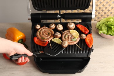 Photo of Woman cooking homemade sausages with bell peppers and mushrooms on electric grill at wooden table, closeup