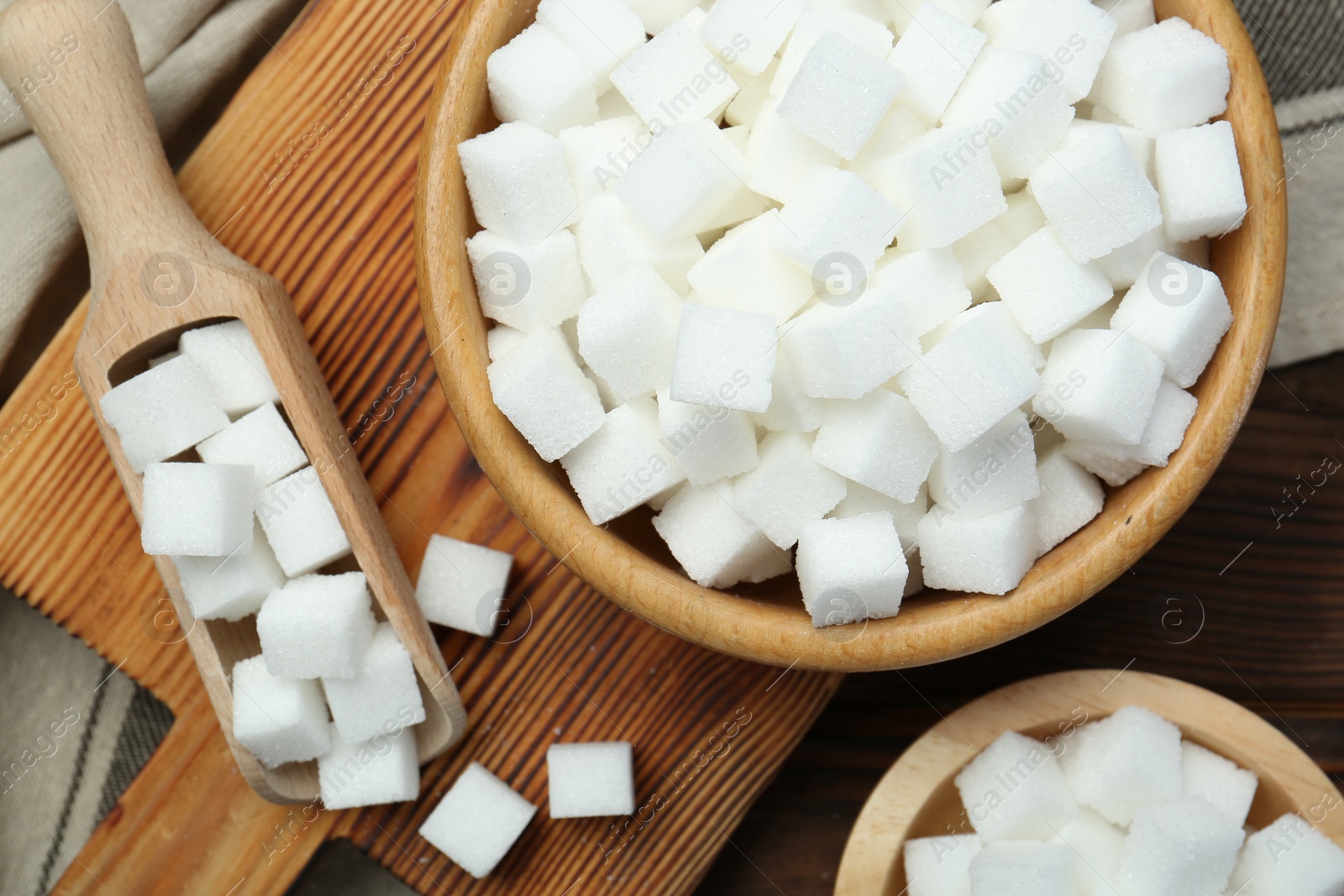 Photo of White sugar cubes in bowls and scoop on wooden table, flat lay