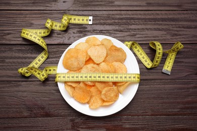 Plate with potato chips and measuring tape on wooden table, flat lay. Weight loss concept