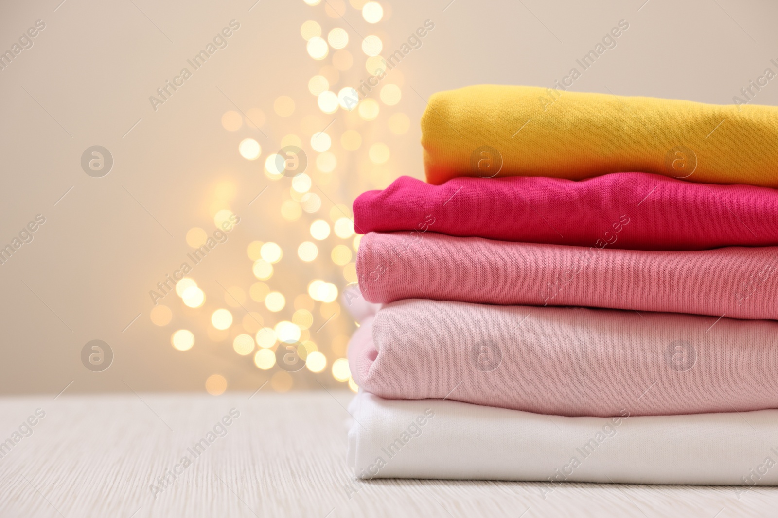 Stack of folded clothes on wooden table against beige background with blurred lights, space for text Photo of Stack of folded clothes on wooden table against beige background with blurred lights, space for text
