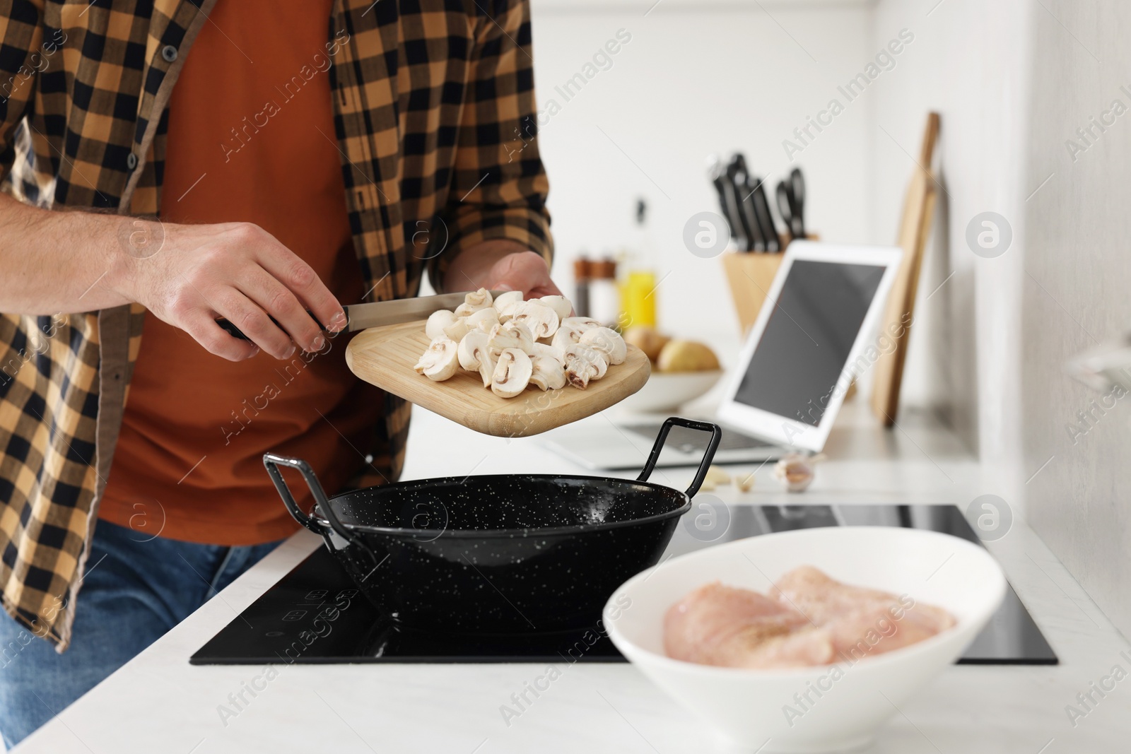 Photo of Man putting cut mushrooms into frying pan while watching online cooking course via laptop in kitchen, closeup