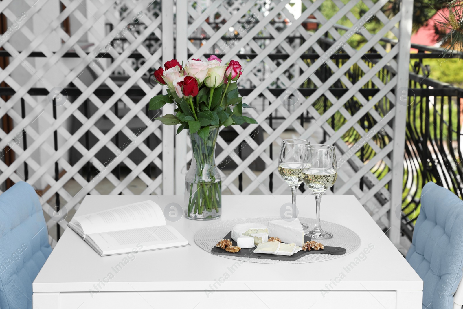 Photo of Vase with roses, open book, glasses of wine and snacks on white table at balcony