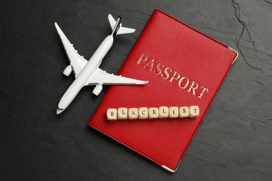 Wooden cubes with word Blacklist, toy airplane and passport on black background, flat lay