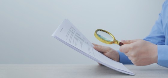 Man looking at documents through magnifier at wooden table, closeup. Banner design with space for text