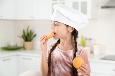 Photo of Cute little girl wearing chef hat eating cookies in kitchen