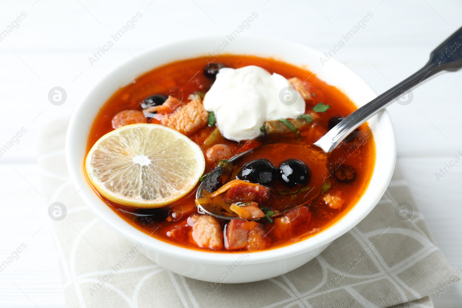 Photo of Meat solyanka soup with sausages, olives, vegetables and spoon on white table, closeup