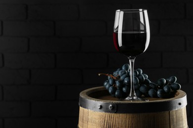 Photo of Glass with delicious wine and fresh grapes on wooden barrel against dark wall, space for text