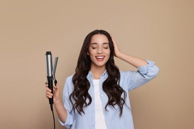 Happy woman with curling hair iron on beige background