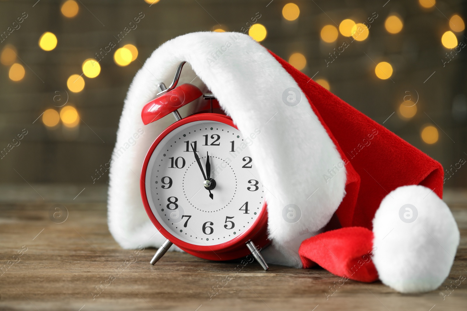 Photo of Alarm clock with Santa hat on wooden table against blurred Christmas lights. New Year countdown