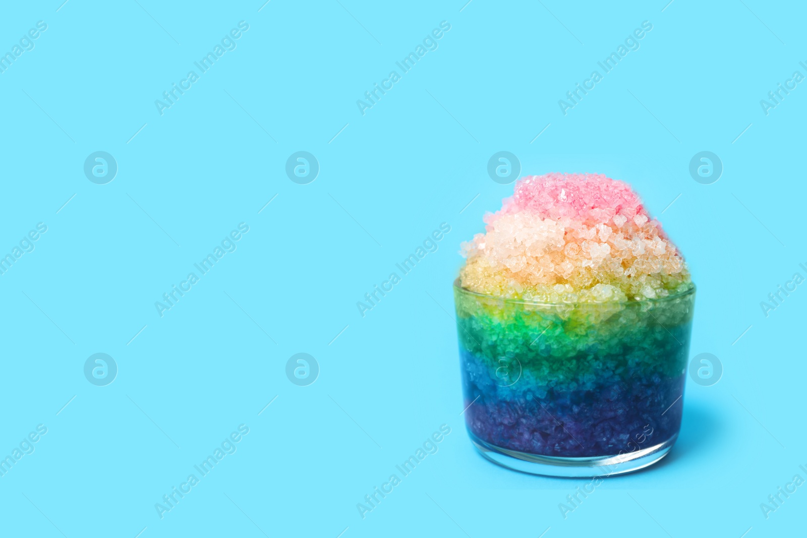 Photo of Rainbow shaving ice in glass dessert bowl on light blue background, space for text