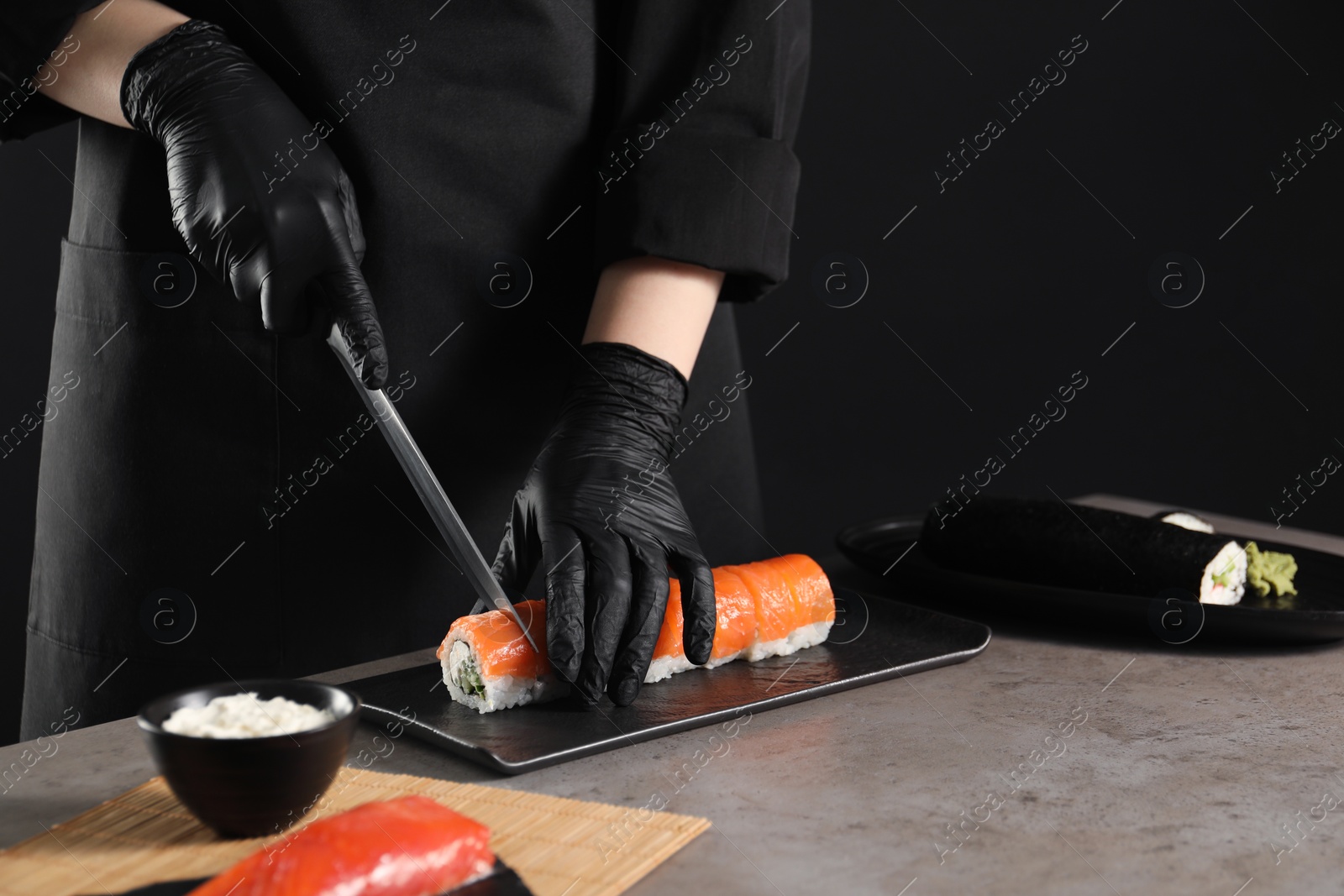 Photo of Chef in gloves cutting sushi roll at grey table, closeup