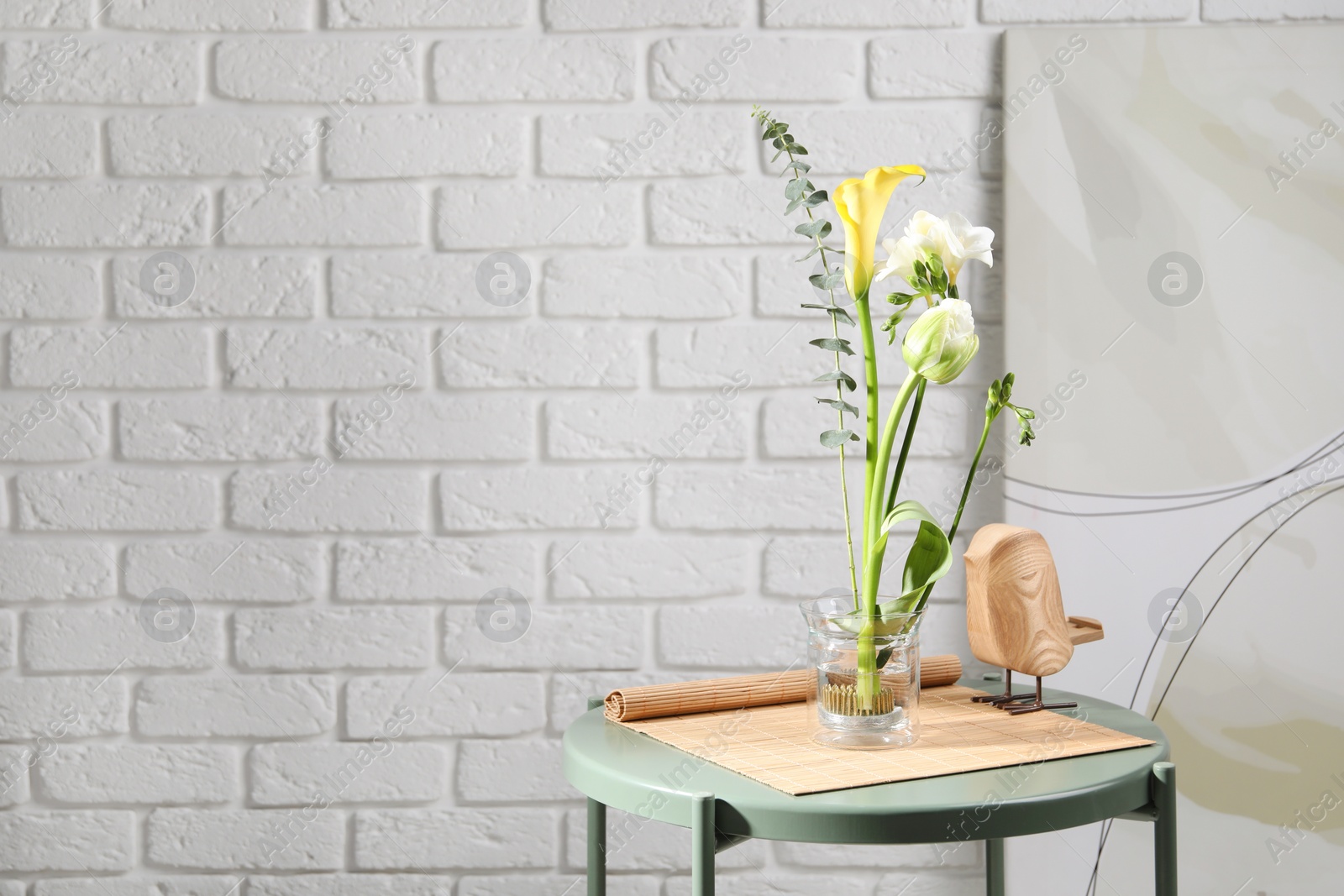 Photo of Beautiful ikebana for stylish house decor. Floral composition with fresh flowers and eucalyptus branch on coffee table near white brick wall, space for text