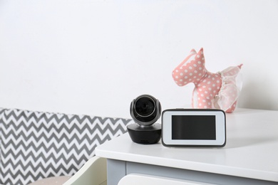 Baby monitor with camera and toy on chest of drawers near crib in room, space for text. Video nanny