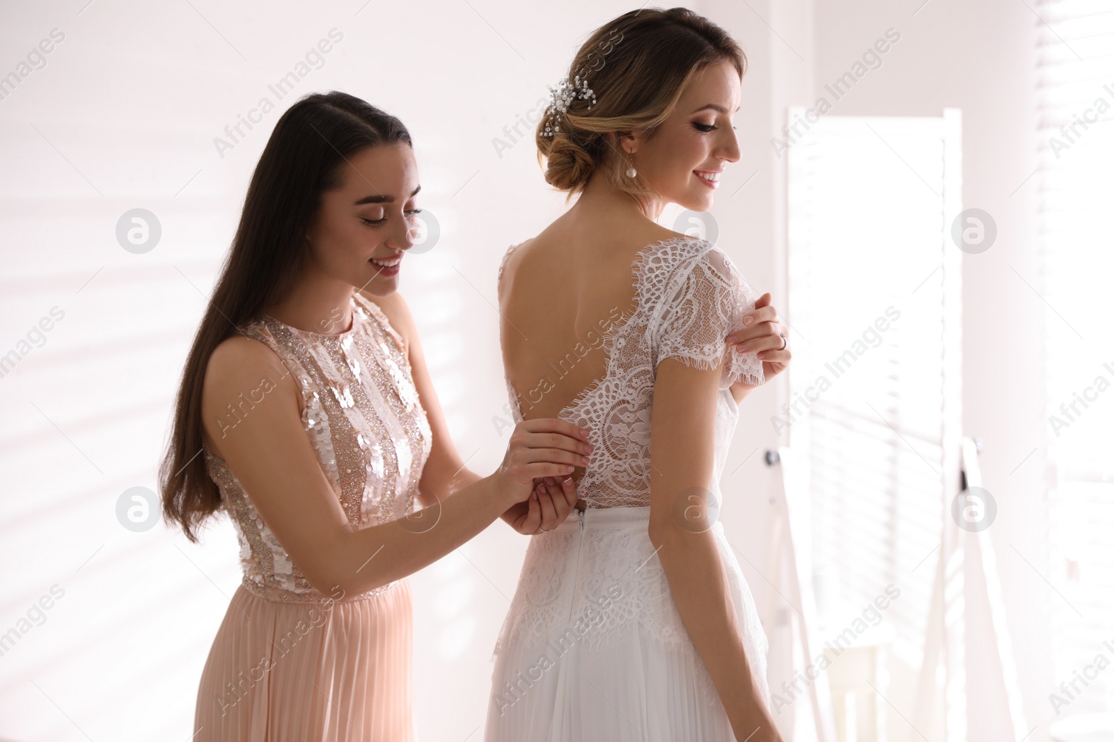 Photo of Young woman helping bride to put on wedding dress in room