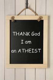 Image of Small chalkboard with phrase Thank God I Am Atheist hanging near white wooden wall