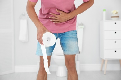 Young man with bath tissue standing near toilet bowl at home