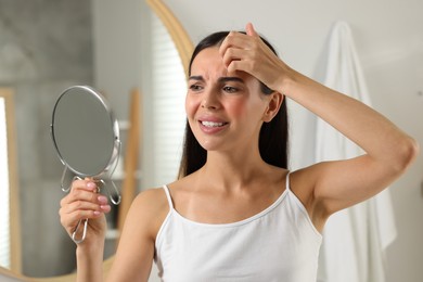 Photo of Suffering from allergy. Young woman looking in mirror and scratching her forehead indoors