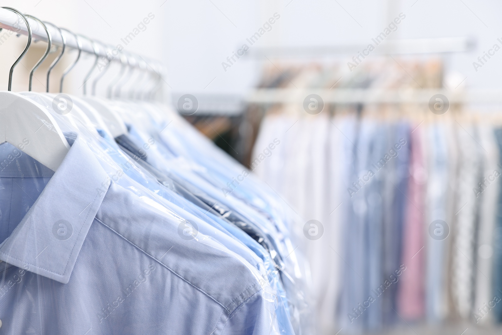 Photo of Dry-cleaning service. Many different clothes in plastic bags hanging on rack indoors, closeup