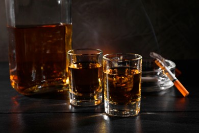 Photo of Alcohol addiction. Whiskey in glasses, smoldering cigarette and ashtray on black wooden table