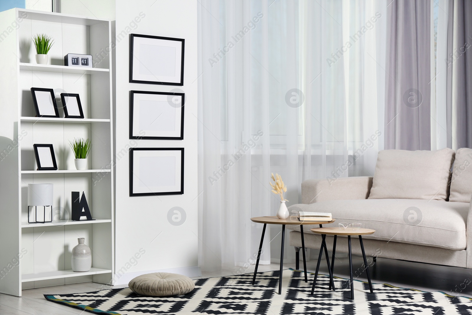 Photo of Stylish room interior with empty frames hanging on white wall near shelving unit