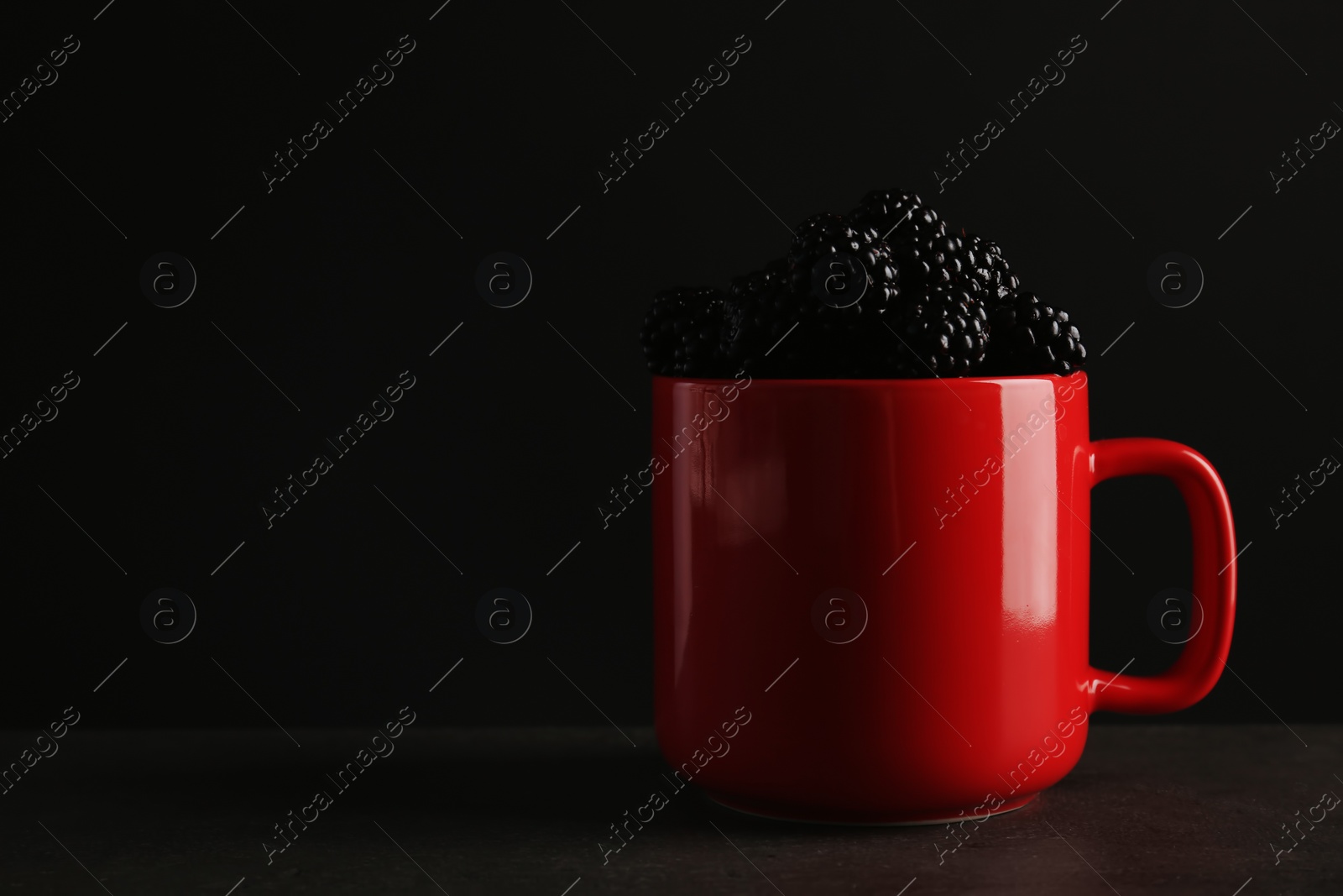 Photo of Mug with fresh blackberry on gray table against dark background. Space for text